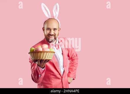 Funny young man in suit and cute bunny ears holding bowl of Easter eggs and smiling Stock Photo
