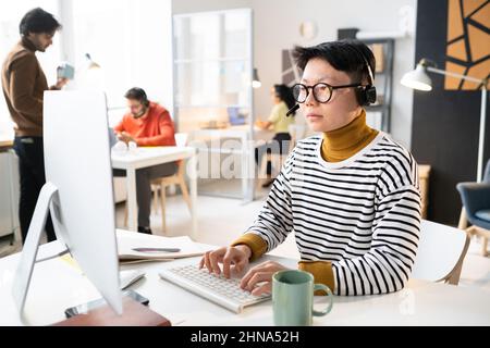 Asian operator in headphones sitting at the table with computer and typing on keyboard, she working with clients online Stock Photo
