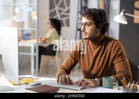 Call centre operator in headphones talking to the customer on the phone and typing on computer keyboard during his work at the table at office Stock Photo