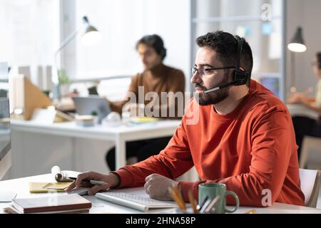 Young bearded man in headphones speaking to the microphone and talking to the clients while using computer at the table at office Stock Photo