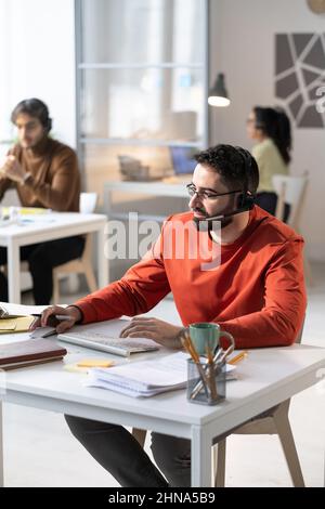 Call center worker in headphone talking to the customers using computer while sitting at the table at office Stock Photo