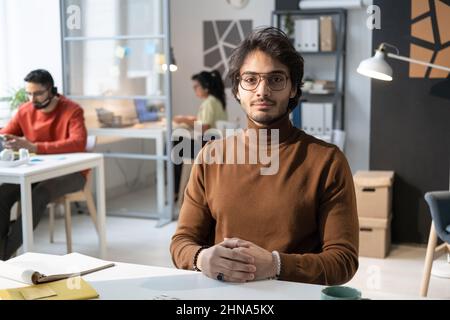 Portrait of young businessman in eyeglasses sitting at his workplace and looking at camera with colleagues in the background Stock Photo