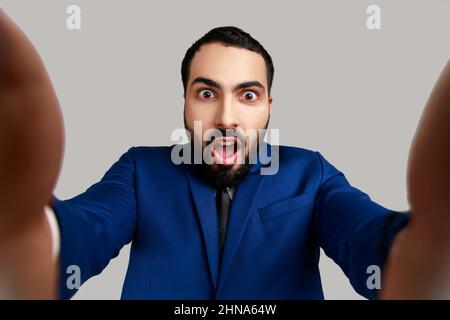 Astonished bearded young adult man taking selfie, looking at camera with shocked expression and open mouth POV, wearing official style suit. Indoor studio shot isolated on gray background. Stock Photo