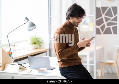 Young businessman leaning on the office table and smiling, he reading a message on his mobile phone Stock Photo