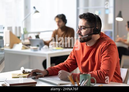 Young bearded man in headphones speaking to the microphone and talking to the clients while using computer at the table at office Stock Photo
