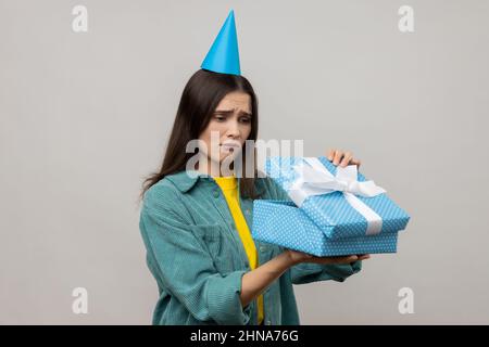 Portrait of upset woman opening birthday gift box and looking inside with disappointed expression, unwrapping bad present, wearing casual style jacket. Indoor studio shot isolated on gray background. Stock Photo