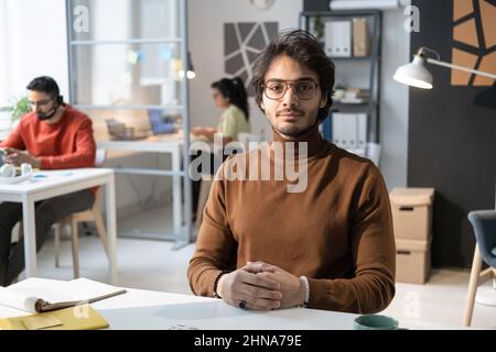 Portrait of young businessman in eyeglasses sitting at his workplace and looking at camera with colleagues in the background Stock Photo