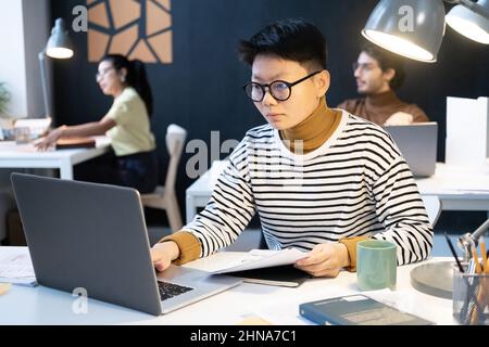 Asian businesswoman in eyeglasses holding documents in front of her and typing on laptop computer while working at her workplace at office Stock Photo