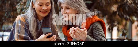 Smiling senior Asian lady shows photos on phone to friend sitting in street cafe Stock Photo