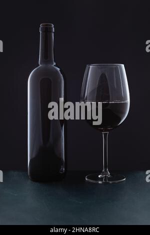 On a green wooden surface, in front of a black background, there is a bottle of red wine without a label and a full glass cup. Stock Photo