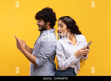 Shocked indian woman spying on her smiling boyfriend who using smartphone, chatting or scrolling social media feed Stock Photo