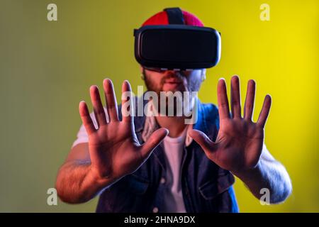 Excited impressed hipster man standing in virtual reality headset, trying to touch something stretching his arms forward, wearing hat. Indoor studio shot isolated on colorful neon light background. Stock Photo