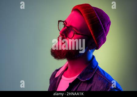 Portrait of handsome bearded hipster man in glasses, looking away with dreamy expression, wearing red beanie hat, beauty and fashion. Indoor studio shot isolated on colorful neon light background. Stock Photo