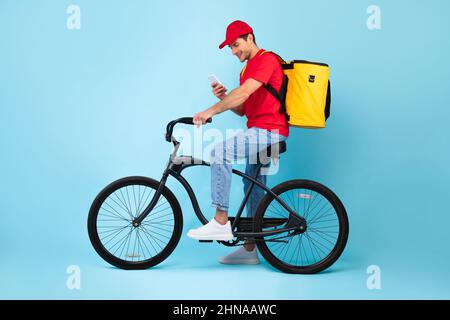 Courier Using Smartphone Sitting On Bike Delivering Food, Blue Background Stock Photo