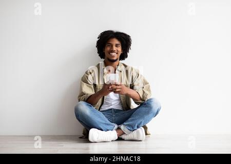 Mobile App. Young black man with smartphone in hands relaxing on floor Stock Photo