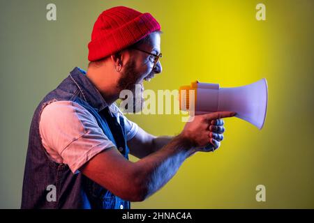 Side view of bearded hipster man yelling holding megaphone, looking to right side, protesting, wearing beanie hat and denim vest. Indoor studio shot isolated on colorful neon light background. Stock Photo