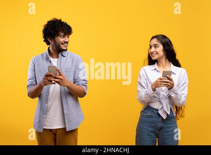 Cheerful young indian man and woman in casual outfits holding smartphones, looking at each other and smiling Stock Photo
