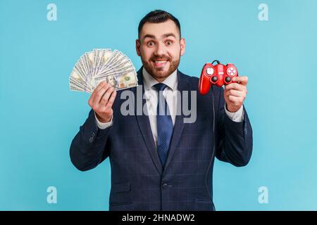 Portrait of smiling bearded businessman wearing dark official style suit holding in hands dollar banknotes and red joystick, looking at camera. Indoor studio shot isolated on blue background. Stock Photo