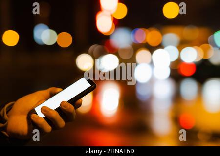 Smartphone with white screen in human hand, copy space in darkness with bright bokeh lights. Using phone in the city at night Stock Photo