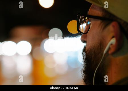 Close-up portrait of a man wearing glasses and earphones in front of bokeh lights at night. Male pedestrian with city street bokeh background, shallow Stock Photo