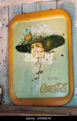 Vintage French language Coca Cola metal tray with silkscreen printed woman's portrait on wall inside old rustic wood plank cabin. Stock Photo