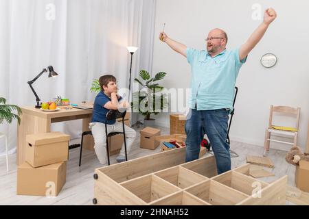 Dad is very happy and son is sitting at his desk watching. Assembling furniture. Stock Photo