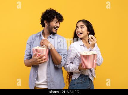Overjoyed indian man and woman eating popcorn, standing back to back and smiling at each other, yellow background Stock Photo