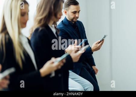 Portrait diverse candidates sitting on chairs in row, looking at camera, business people applicants interns waiting for job interview, holding gadgets Stock Photo