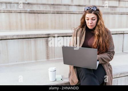 Woman in outerwear browsing data on netbook while sitting on concrete stairs near smartphone and drink to go in daytime in city Stock Photo