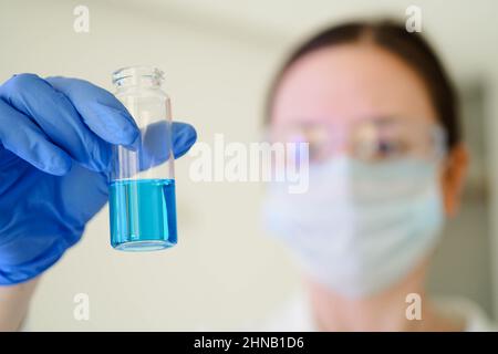 Serious female scientific researcher in gloves looking at a test tube with blue liquid in a laboratory. Close up. Stock Photo