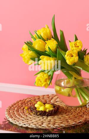 Vase with tulips and nest with Easter eggs on table near color wall Stock Photo