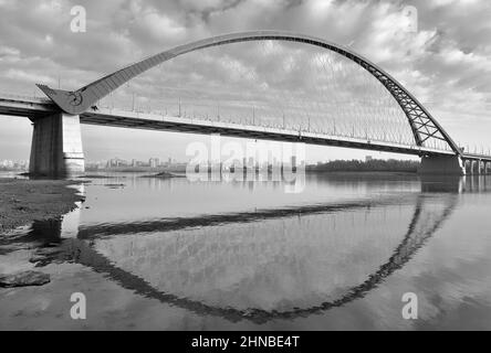 The bank of the Ob River. Bugrinsky arch bridge with reflection in the water on the outskirts of a big city. Novosibirsk, Siberia, Russia, 2021 Stock Photo