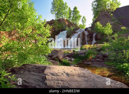 Belovsky waterfall in the spring. A small waterfall among the rocks, surrounded by birch trees with spring foliage in the rays of the sun. White strea Stock Photo