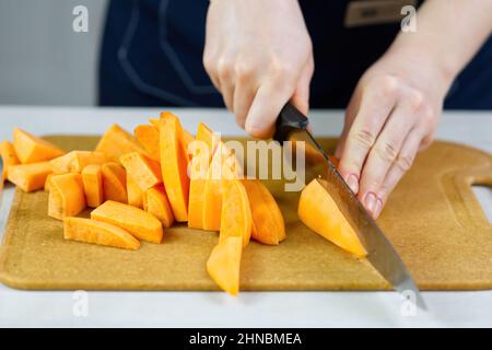 woman cutting with knife sweet potato into wedges on wooden board. Stock Photo