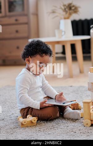 Concentrated curly black boy sitting on carpet in living room and watching cartoons on tablet Stock Photo