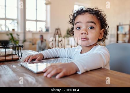 Portrait of surprised curious African-American boy with curly hair sitting at table with gadget Stock Photo