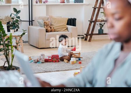 African-American toddler sitting on carpet and playing with toys alone while his mother working, focus in background Stock Photo