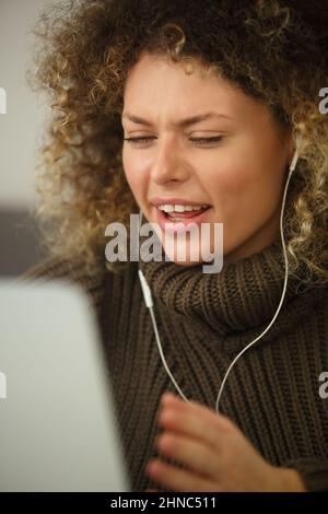 Emotional curly woman talking on video call. Fun white female with diastem and curls speaking on headset and laptop web camera Stock Photo