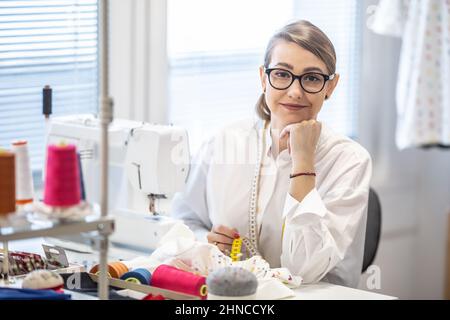 Good-looking demale tailor wears glasses sitting behind the desk, smiling. Stock Photo