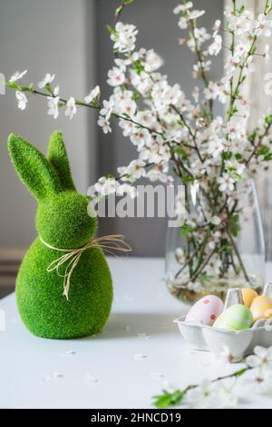 Green easter bunny with blooming flowers on tree branch in the vase and colored easter eggs in container on the white table. Happy Easter spring verti Stock Photo