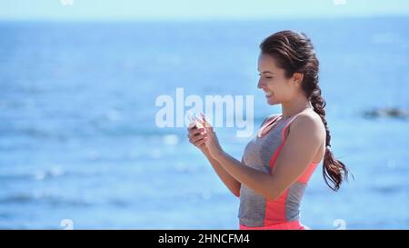 Smiling sportswoman using smartphone near blurred sea Stock Photo