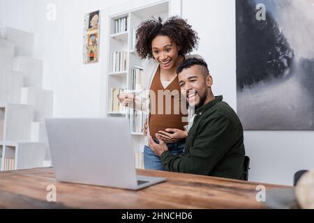 happy pregnant african american woman pointing at belly near husband during video call on laptop Stock Photo