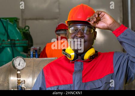 Black Industrial Worker In Red Helmet, Protective Eyewear, Hearing Protection Equpiment And Work Uniform. Stock Photo
