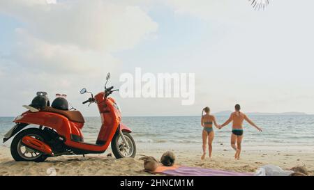 Scooter road trip. Lovely couple on red motorbike in white clothes on sand beach. Just married people kiss hugs walking near the tropical palm trees, sea. Wedding honeymoon by ocean. Motorcycle rent. Stock Photo