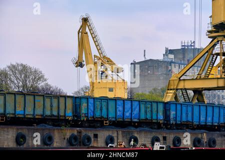 Close view of river port crane loading open-top gondola cars of freight train on cloudy day. Empty river drag boats or barges moored by pier Stock Photo