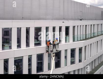 Two workers wearing safety harness wash office building facade at height standing in a crane cradle or aerial platform using pressure washer and mops Stock Photo