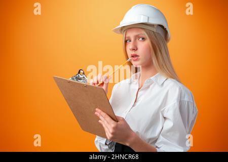Teen girl in hardhat and clipboard against orange background Stock Photo