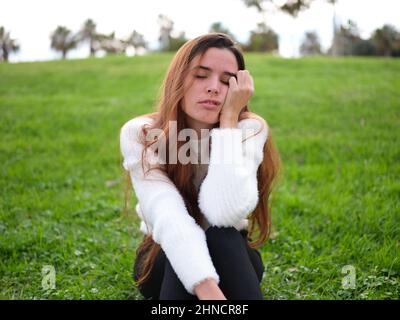 A young woman in the park sitting on the grass with one hand on her face falling asleep. Stock Photo