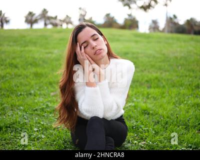 A young woman in the park sitting on the grass with her hands on her face and her eyes closed daydreaming. Stock Photo