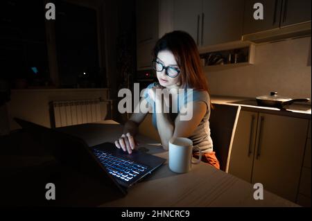 Caucasian woman wearing glasses at a wireless computer at night in the kitchen. Girl works overtime in the home office. Freelancer is typing on a Stock Photo